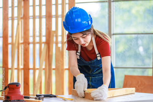 Asian professional female engineer architect foreman labor worker wears safety hard helmet jeans apron and gloves standing using squard angle degree ruler and pencil measuring wood plank on workbench photo