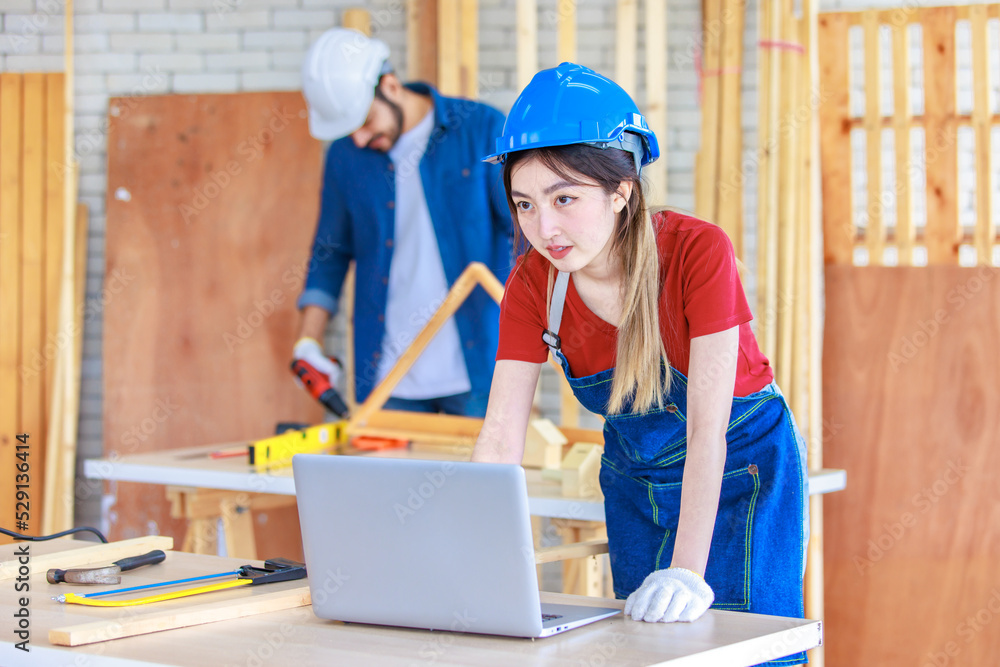 Asian professional female engineer architect foreman labor worker wears safety goggles and jeans apron standing smiling using laptop computer working on wooden table with tools in construction site Asian professional female engineer architect foreman labor worker wears safety goggles and jeans apron standing smiling using laptop computer working on wooden table with tools in construction site