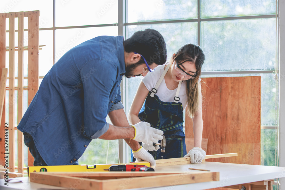 Asian young female engineer architect foreman labor worker wears safety goggles gloves and apron holdind using steel hammer pounding nail on wood stick when Indian professional male carpenter helping Asian young female engineer architect foreman labor worker wears safety goggles gloves and apron holdind using steel hammer pounding nail on wood stick when Indian professional male carpenter helping