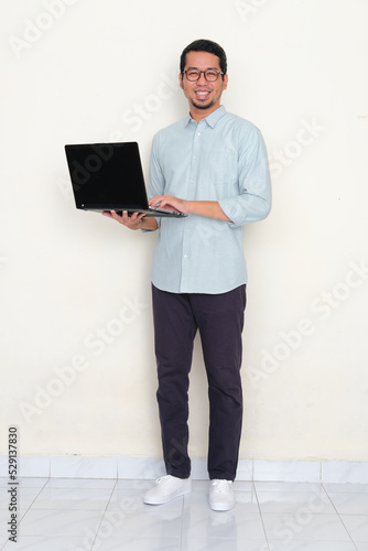 Full body portrait of Asian man smiling to camera while holding a laptop photo