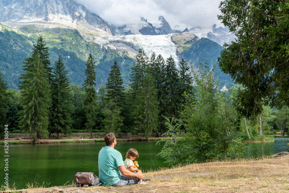 Father and his little daughter sitting on a lake shore looking at Mont Blanc slopes in Chamonix, France. Father and his little daughter sitting on a lake shore looking at Mont Blanc slopes in Chamonix, France.