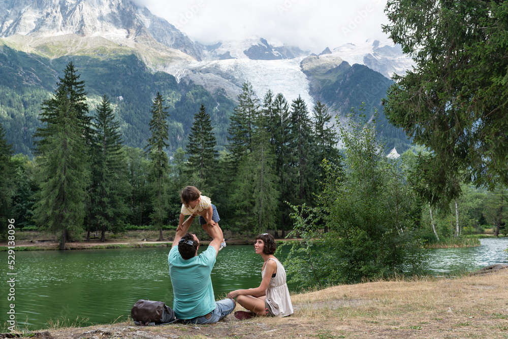 Father holding her little daughter up while mother is looking at them, both sitting on a shore lake with the majestic Montblanc on the background.