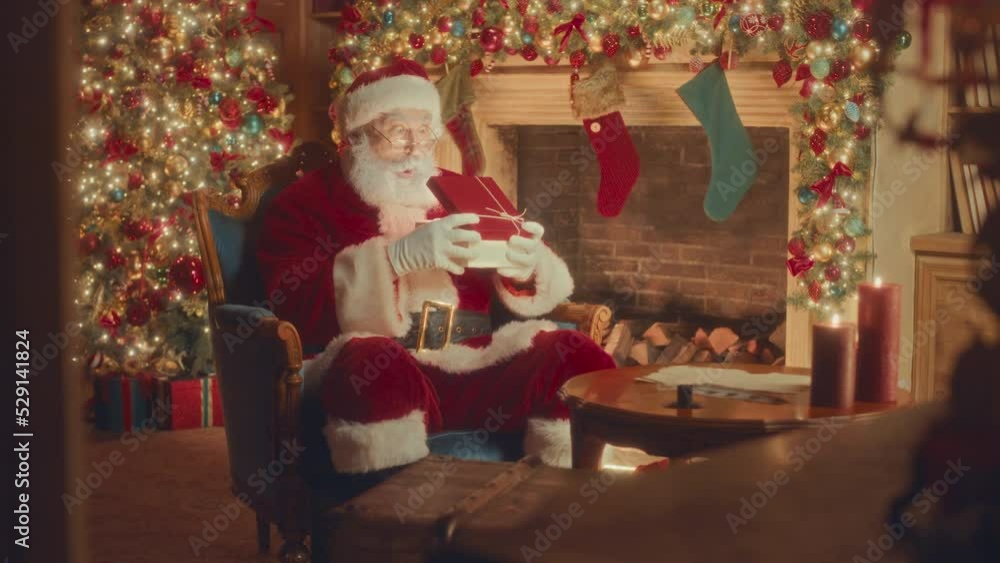 Slowmo of surprised Santa Claus in red costume opening Christmas present box sitting by beautiful fireplace decorated with sparkling lights Slowmo of surprised Santa Claus in red costume opening Christmas present box sitting by beautiful fireplace decorated with sparkling lights