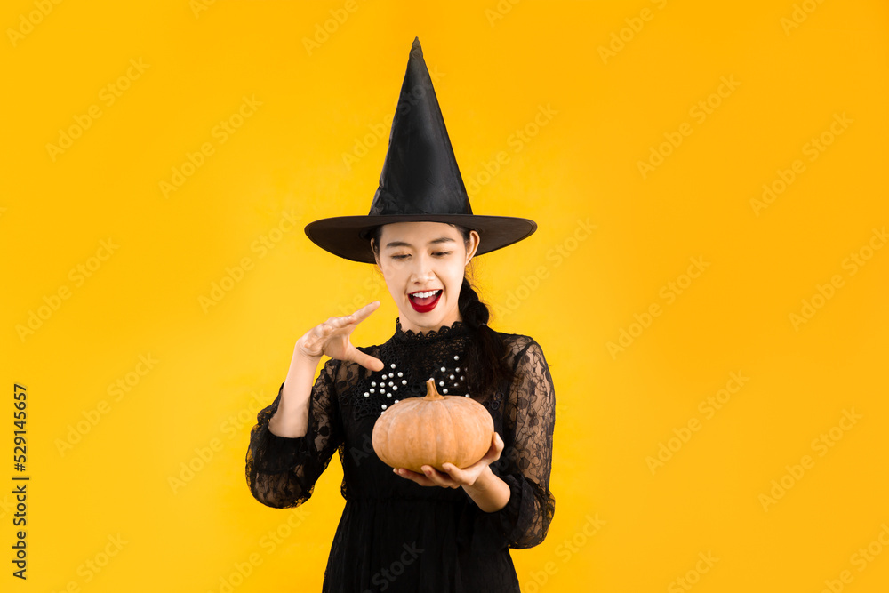 Halloween theme, young asian woman in witch costume black dress wearing hat holding pumpkin on yellow color background.