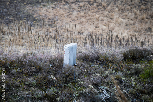 Canvas-taulu An electrical heater dumped in grassland with dry bushes and wired fence