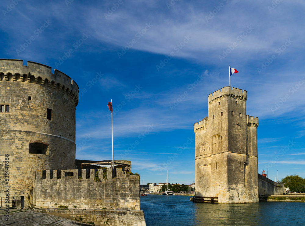 View of the entrance to the old port of the French city of La Rochelle with the two medieval towers and blue sky with light clouds.