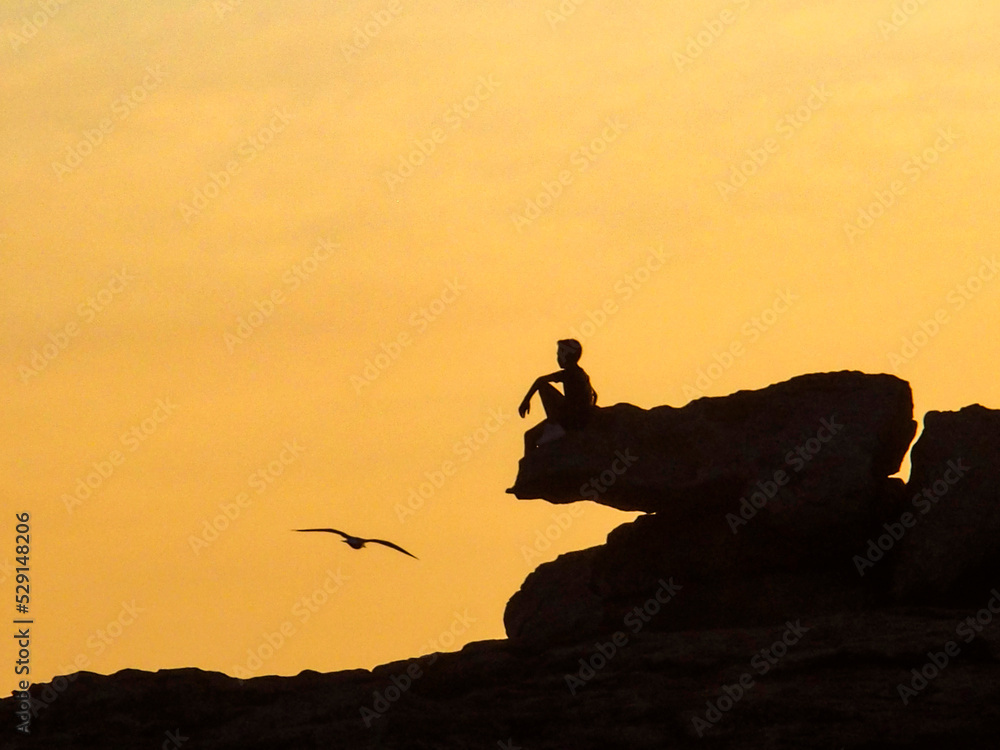 Silueta de un niño sentado en una roca con el fondo naranja del atardecer. Una gaviota aparece en la escena. Castro de Baroña, Galicia, España.