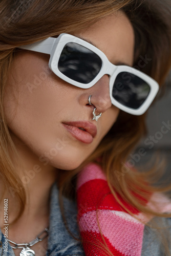 Closeup fashion female portrait of a stylish pretty young hipster girl with nose piercing with fashionable white sunglasses wearing a denim jacket and pink sweatshirt stands on the street