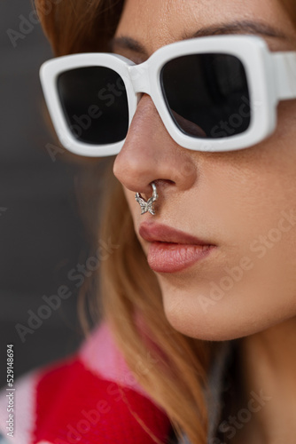 Amazing street closeup portrait of pretty fashion woman with stylish white sunglasses with noise piercing in fashionable urban clothes on a dark background. Female hipster face