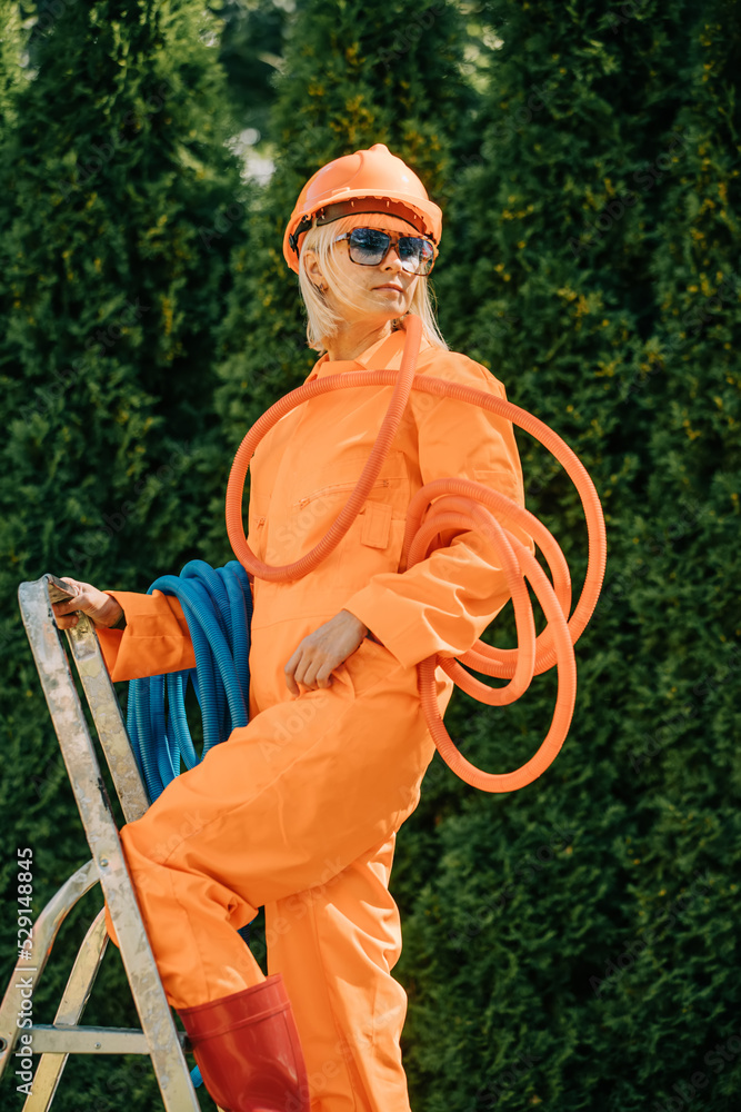 Stylish woman in orange suit, hardhat, pipes and sunglasses next to stepladder