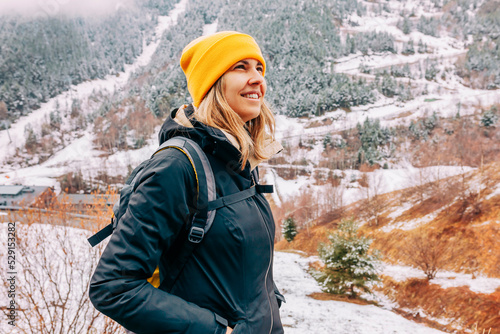 Winter hike on snow mountain young happy hiker woman climbing. Europe travel adventure trek in nature landscape. Young cheerful female wearing yellow hat, black jacket for cold weather and bag.