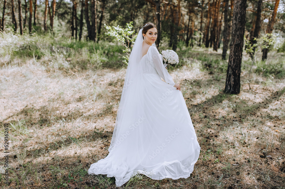 A beautiful brunette bride in a white long dress with a bouquet of roses stands in a coniferous forest in nature. Wedding portrait, photography.