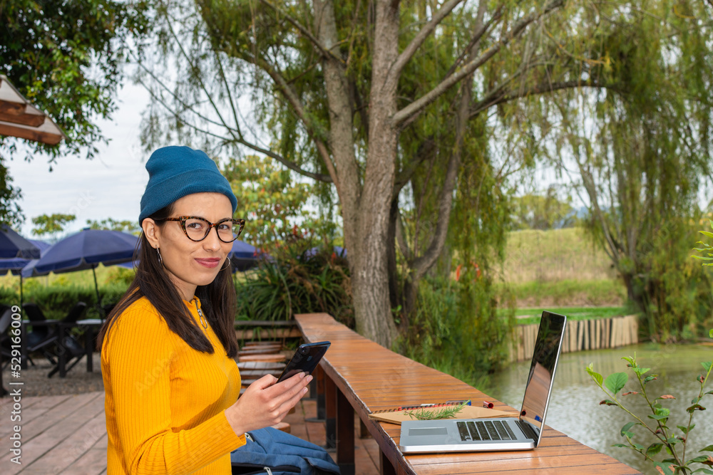 Latin female student wearing yellow sweater and blue beanie sitting outside holding her cell phone while looking straight at the camera Latin female student wearing yellow sweater and blue beanie sitting outside holding her cell phone while looking straight at the camera