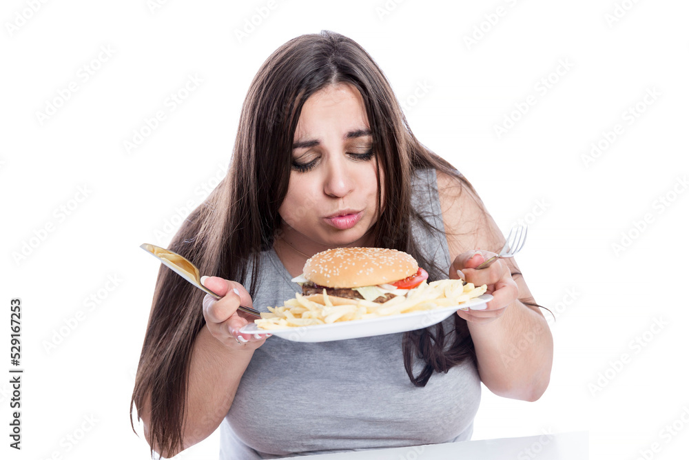An overweight woman looks with appetite at a plate of french fries and a hamburger. Junk food and culture. Brunette in a gray shirt. Isolated on white background. An overweight woman looks with appetite at a plate of french fries and a hamburger. Junk food and culture. Brunette in a gray shirt. Isolated on white background.