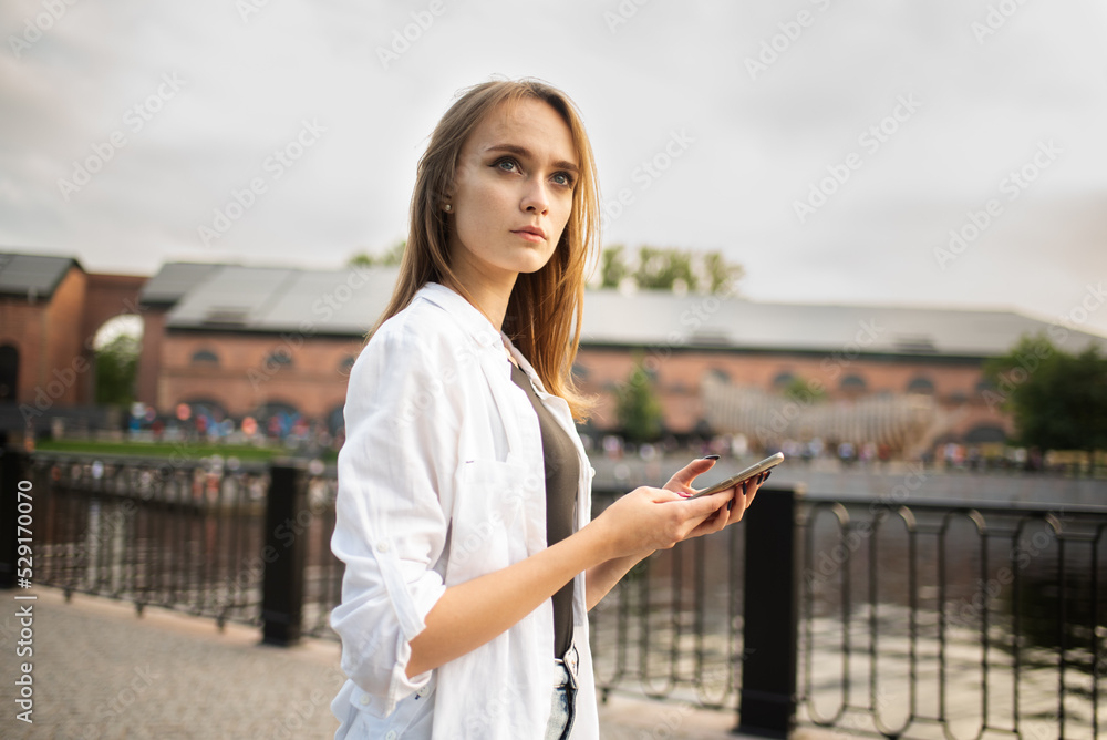 A young and attractive Caucasian girl in casual clothes uses a mobile phone outdoors.