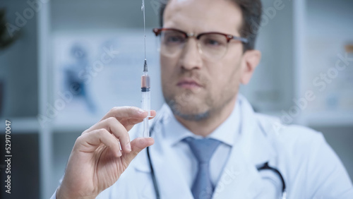 blurred doctor in white coat and glasses holding syringe in clinic.