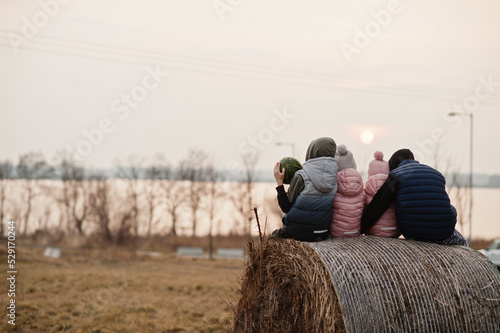 Back of four kids sitting on haycock at field against lake sunset. photo