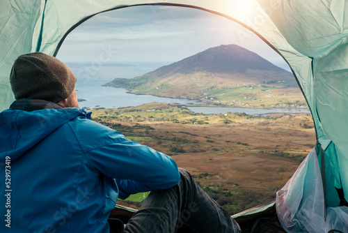 Male tourist sitting in a tent and looking out at stunning nature landscape with ocean and mountains Travel in nature concept. Model in grey hat and blue jacket and jeans. Diamond hill, Ireland.