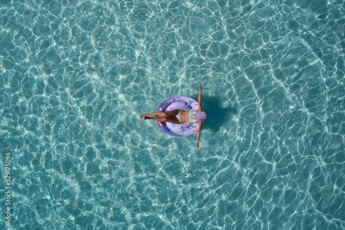 Girl in a turquoise swimsuit in water color, pool ring purple on transparent turquoise water top view. Girl relaxing vacation in the pool top view. Woman on turquoise water aerial view.