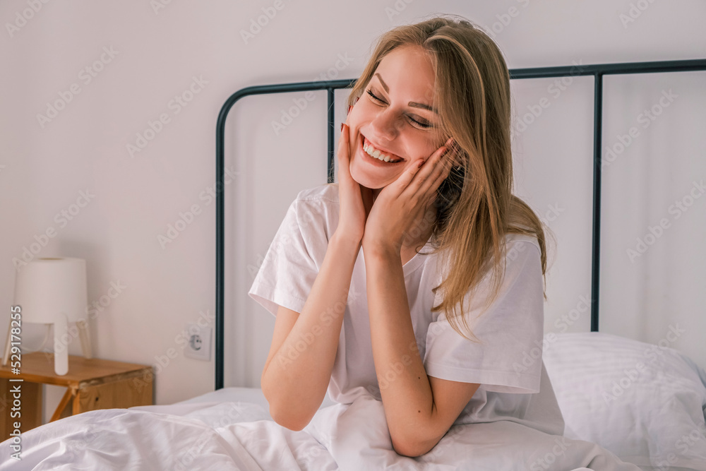 Waking up in the morning. A young happy woman smiling and touching her face sitting in bed after waking up. Morning bliss. Healthy sleep, body recovery Waking up in the morning. A young happy woman smiling and touching her face sitting in bed after waking up. Morning bliss. Healthy sleep, body recovery