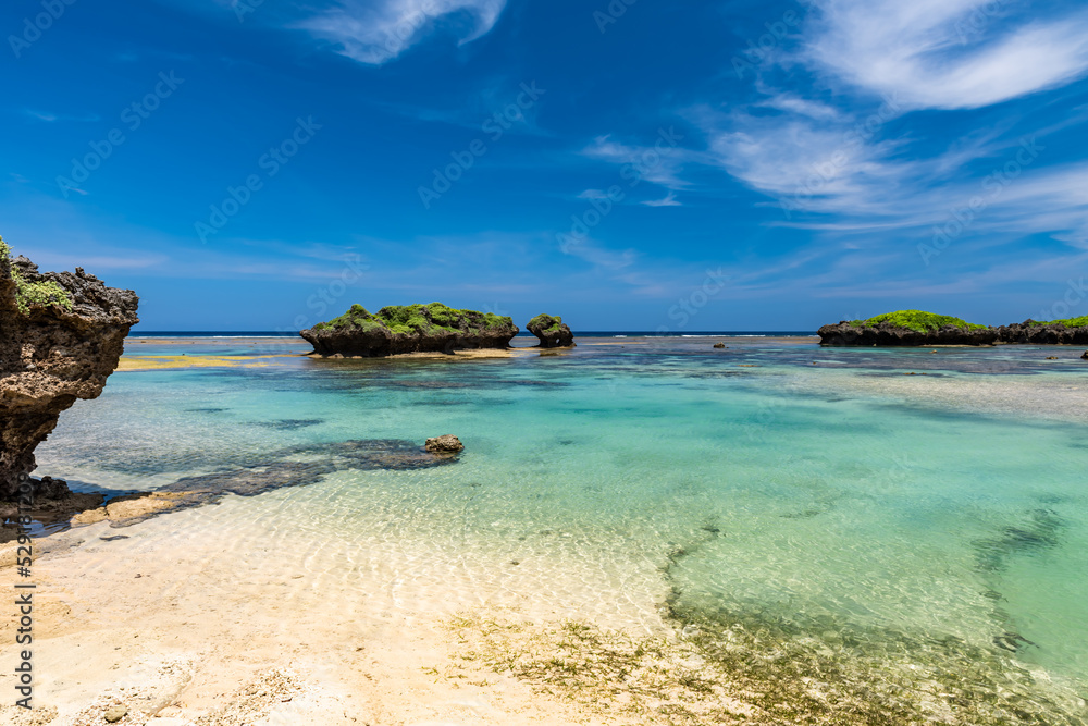 Stunning crystal clear turquoise sea waters forming natural pools between coral rocks, green islets, blue sky.