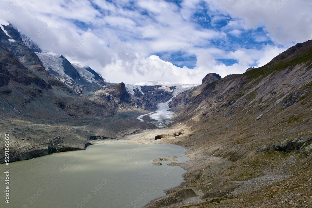 Scenic view of Pasterze Glacier and the lake bellow in Glockner Group of the High Tauern mountain range in Carinthia, Austria with clouds covering the mountain peaks and blue sky above