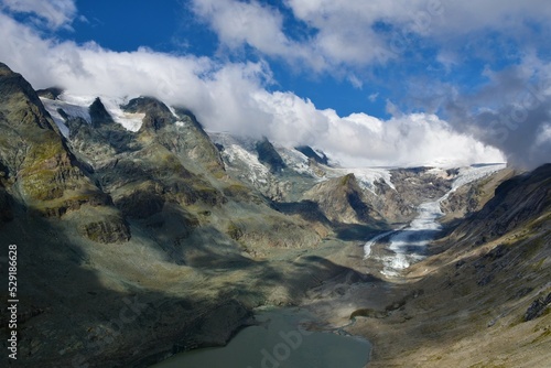 Scenic view of Pasterze Glacier and mountains of the Glockner Group in High Tauern in Austrian Central Alps, Carinthia, Austria with the peaks in clouds and blue sky above © kato08