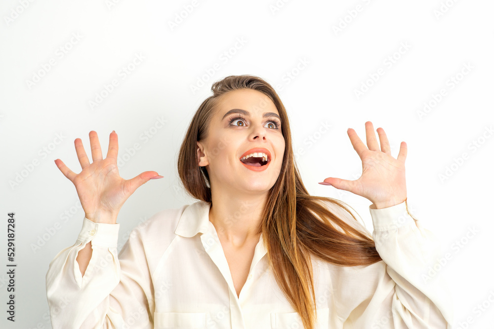 Portrait of young caucasian woman wearing white shirt raises hands and laughs positively with open mouth looking up against a white background Portrait of young caucasian woman wearing white shirt raises hands and laughs positively with open mouth looking up against a white background