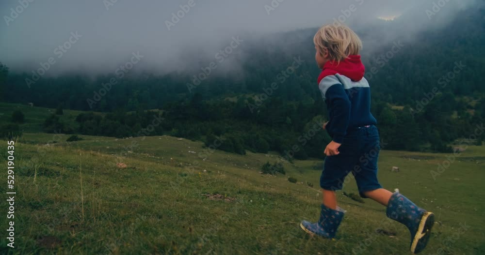 Joyful child boy run in rubber boots on rural environment at autumn hike travel onto mountain feeling free. Low clouds strew over mountain peak. Carefree childhood on countryside active leisure Joyful child boy run in rubber boots on rural environment at autumn hike travel onto mountain feeling free. Low clouds strew over mountain peak. Carefree childhood on countryside active leisure