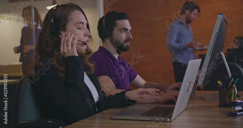 Telemarketers communicating with clients wearing headsets in front of laptops. Female and male employees working inside office workplace speaking with customer in call center support