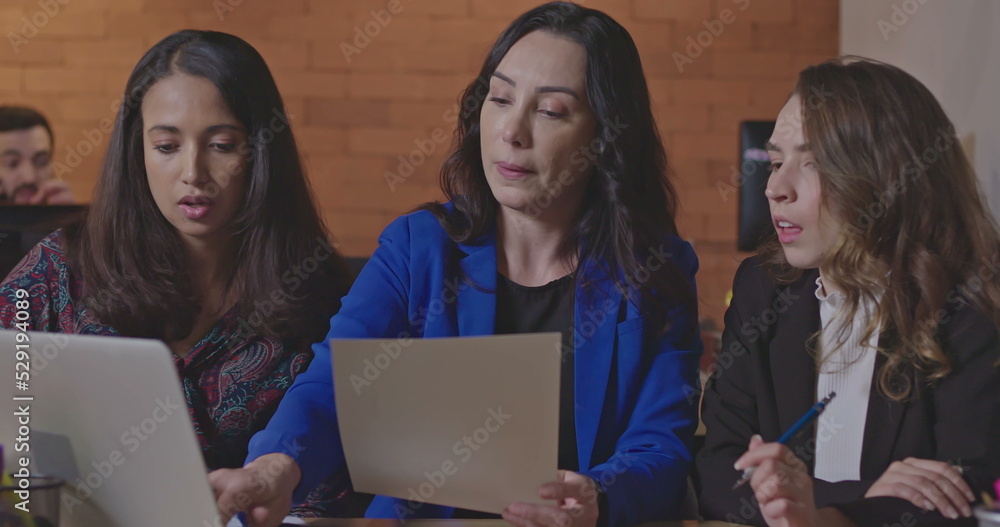 Women discussing work in front of laptop computer at corporate workplace. Female leader coaching younger female staff. People collaborating at night in the office Women discussing work in front of laptop computer at corporate workplace. Female leader coaching younger female staff. People collaborating at night in the office