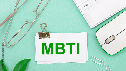 On a light green background there is a white calculator, a computer mouse, green leaves of a plant, gold-rimmed glasses and a white card with text MBTI. Business concept photo