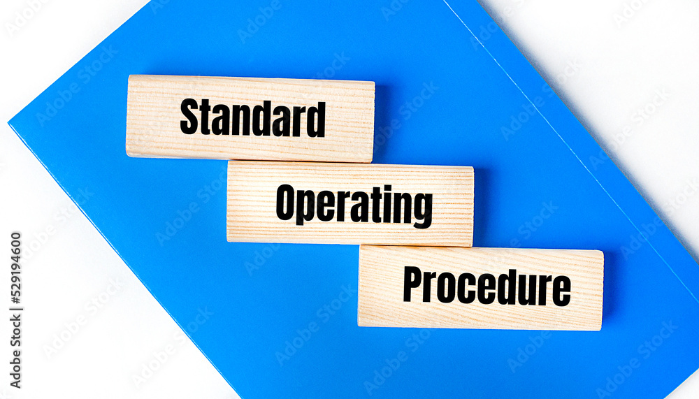 There is a blue notebook on a light gray background. Above are three wooden blocks with the words SOP Standard Operating Procedure There is a blue notebook on a light gray background. Above are three wooden blocks with the words SOP Standard Operating Procedure