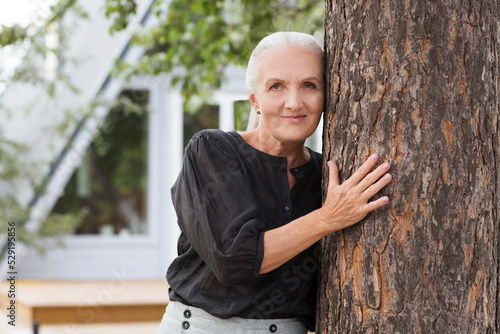mature caucasian woman resting stands by a pine tree near its home. mental healing. copy space. Slow life. Enjoying the little things. spends time in nature in summer