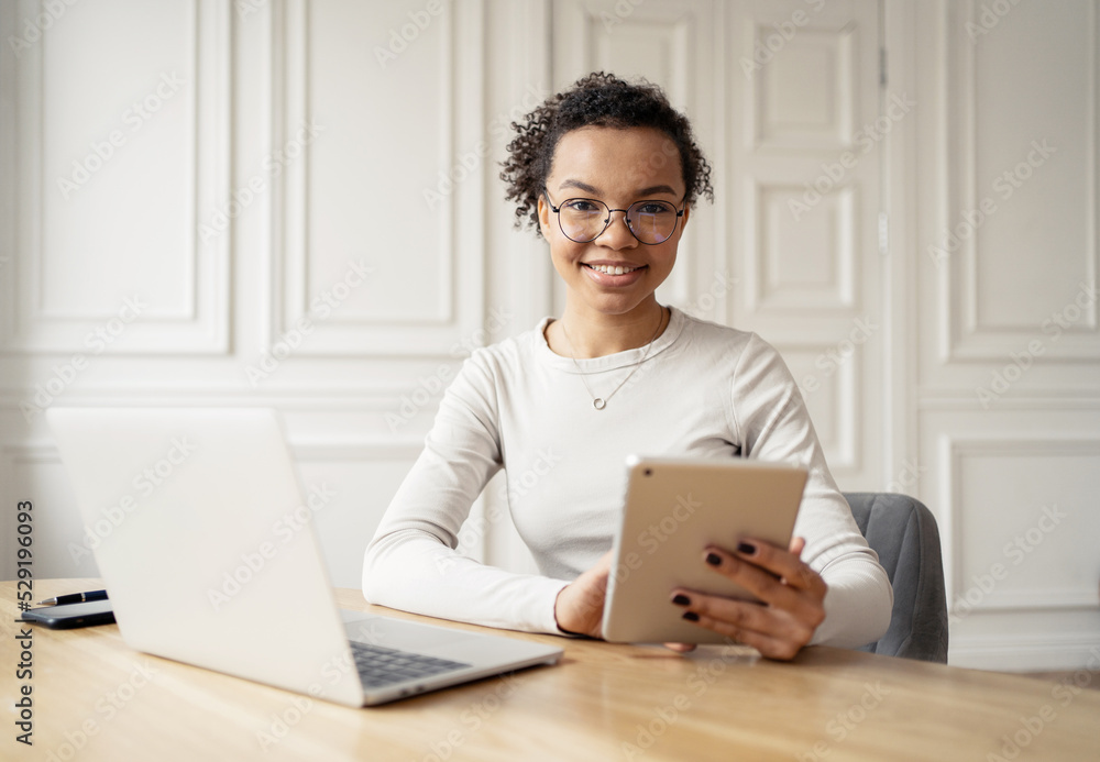 A female freelancer smiles at a video call, uses a laptop and tablet at the workplace in the office A female freelancer smiles at a video call, uses a laptop and tablet at the workplace in the office