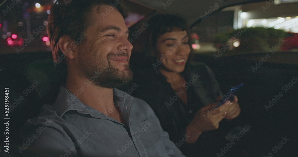Fototapeta premium Young man and woman riding taxi in backseat after work at night. Male passenger looking out car window staring at nightlife city street. People riding cab