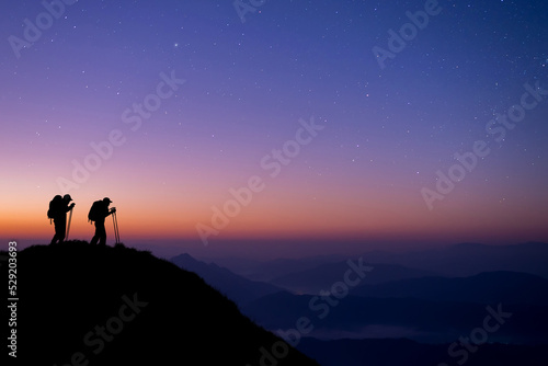 Silhouette of two young hikers were standing at the top of the mountain looking at the stars and Milky Way over the twilight sky. Both of them were happy and free to travel.