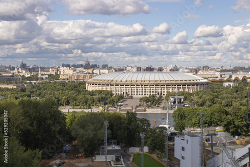 Luzhniki sports stadium, green area with trees and bushes, building constructions and cable car across the river. Cityscape of Moscow. Cloudy sky on a summer day.