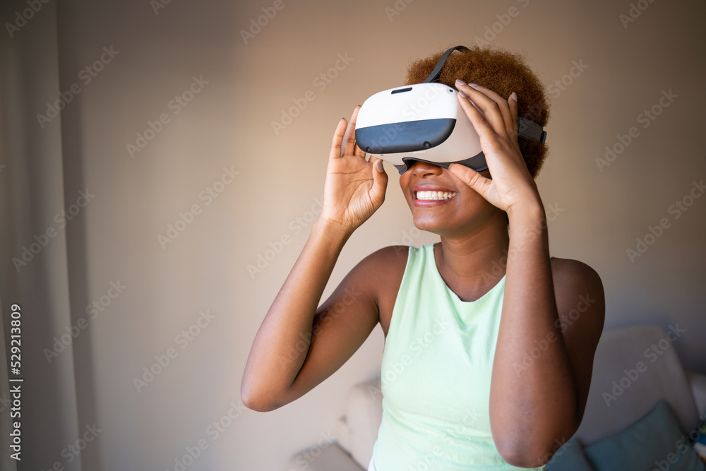 Black skin young woman wearing virtual reality simulator siting on sofa in living room at home. Happy smiling positive emotions. green clothes. touches glasses with hands. Horizontal composition