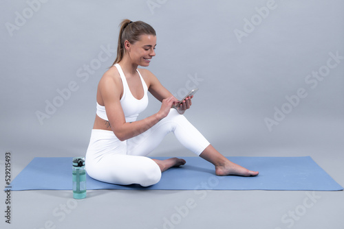 Happy sportswoman using mobile phone. Sportive girl  during exercises on a yoga mat  gray background with space.
