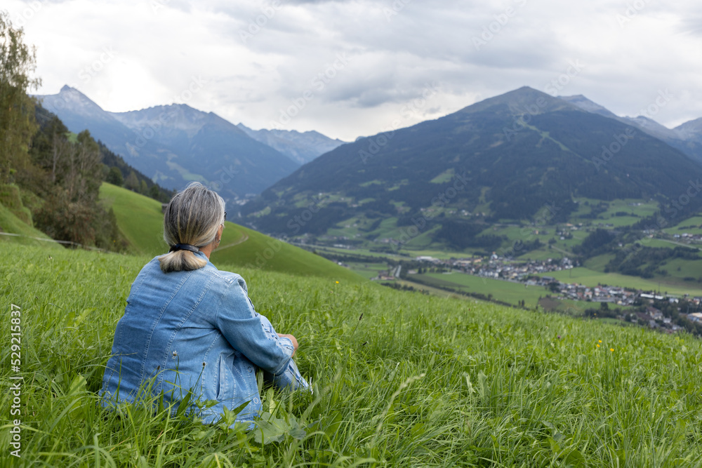 A gray-haired woman in a denim suit sits on a grassy slope and looks towards the mountains.