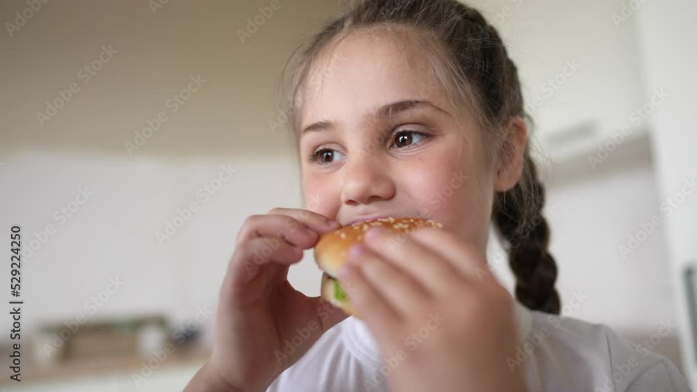 little kid girl eating a hamburger. unhealthy fast food meal proper nutrition concept. child lifestyle greedily with pleasure bites a big burger in the kitchen at home. kid eats fast food close-up little kid girl eating a hamburger. unhealthy fast food meal proper nutrition concept. child lifestyle greedily with pleasure bites a big burger in the kitchen at home. kid eats fast food close-up