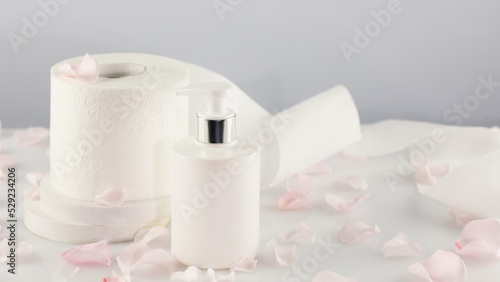 Roll of toilet paper with cosmetic dispenser bottle on light background with rose petals and copy space. White toilet tissue, Hygiene product. Restroom soft touch toilet paper. Soft focus style image
