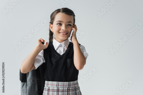happy schoolgirl holding backpack and talking on smartphone isolated on grey. photo