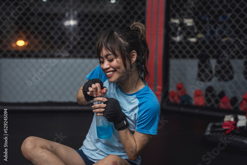 After a demanding training session, a smiling and happy female MMA novice sits down on the ring cage floor and hydrates herself with an electrolyte drink. photo