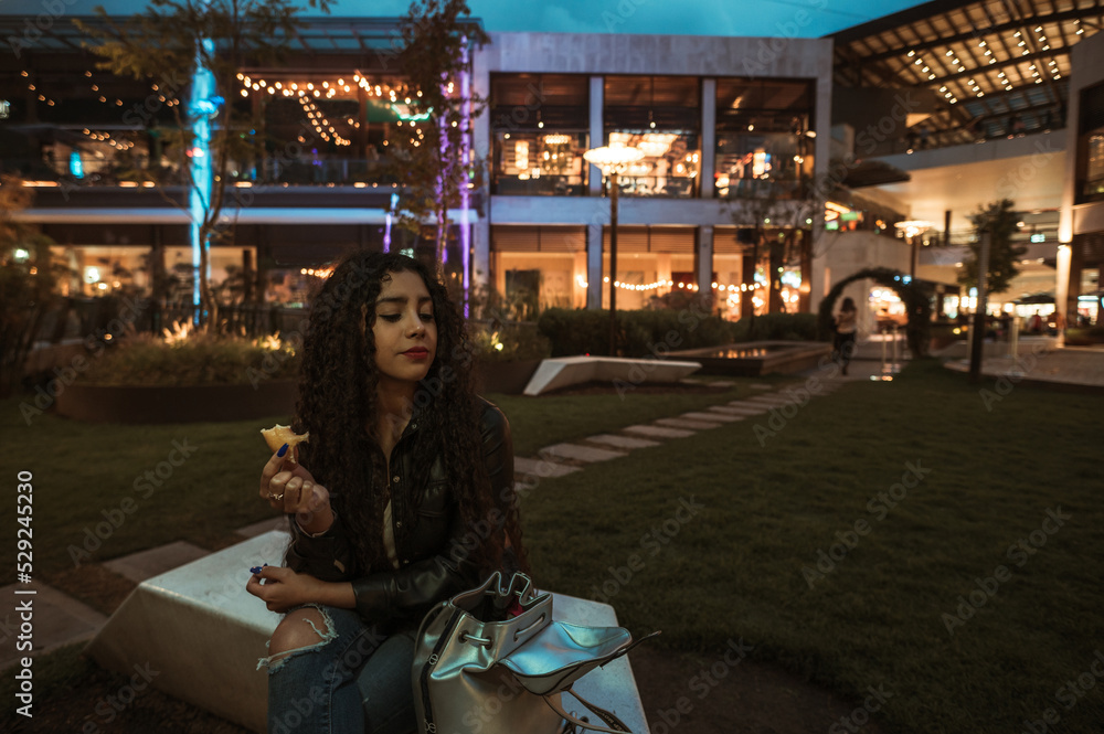 Brunette girl with curly hair enjoys a donut in a place full of lights at nightfall, she wears a black leather jacket. Brunette girl with curly hair enjoys a donut in a place full of lights at nightfall, she wears a black leather jacket.