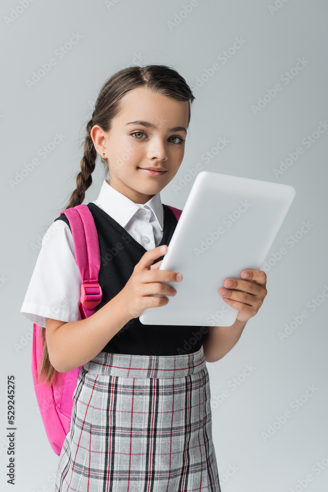 happy and cute schoolgirl in uniform holding digital tablet while smiling isolated on grey. happy and cute schoolgirl in uniform holding digital tablet while smiling isolated on grey.