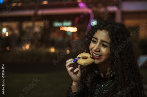 Brunette girl with curly hair enjoys a donut in a place full of lights at nightfall, she wears a black leather jacket.