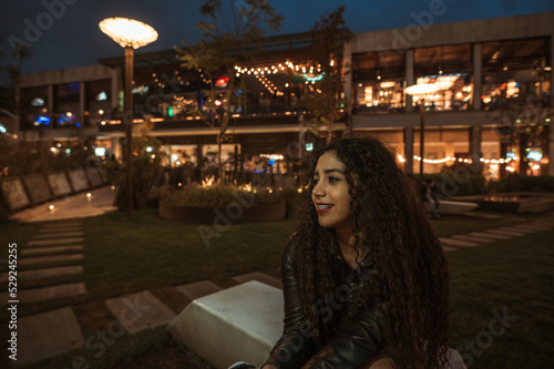 Brunette girl with curly hair enjoys a moment of lights while artificial as night falls, she wears a black leather jacket. © Marco