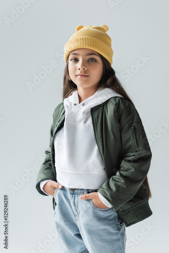 portrait of adorable girl in yellow beanie hat and stylish autumnal outfit posing with hands in pockets isolated on grey.